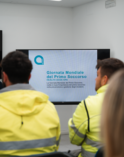 Due dipendenti Acea ATO 5 con giacche alta visibilità seguono la slide di apertura della Giornata Mondiale del Primo Soccorso.