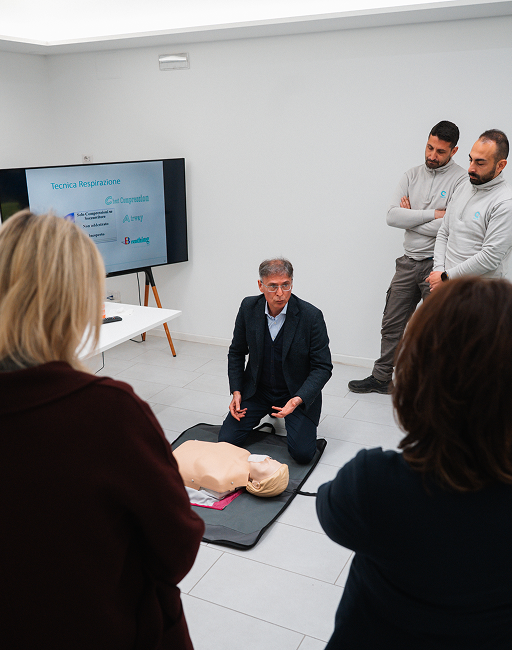 Medico inginocchiato accanto a un manichino spiega la tecnica di rianimazione cardiopolmonare ai partecipanti raccolti intorno a lui.