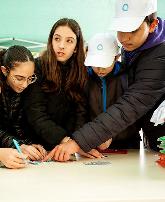 Quattro giovani che indossano giacche invernali e cappellini bianchi lavorano insieme attorno a un tavolo, chinandosi per collaborare a quello che sembra essere un progetto o attività pratica. Una persona a sinistra porta gli occhiali e tiene uno strumento di scrittura blu, mentre gli altri si avvicinano per osservare e partecipare all'attività.